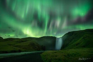 Northern lights of green, white and pink dance in the sky over a lush green landscape where a waterfall tumbles over a cliff in the distance. 