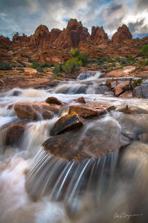 Water cascades down the red desert rocks from the distant rock formations with a blue sky and white clouds in the background.