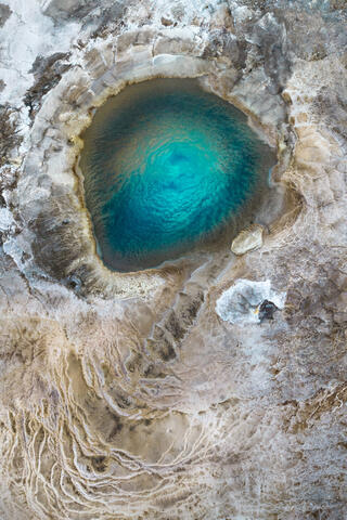 Aerial photograph of a geyser filled with bright blue water and surrounded by light tan rock creates an abstract image of what looks like a portal.