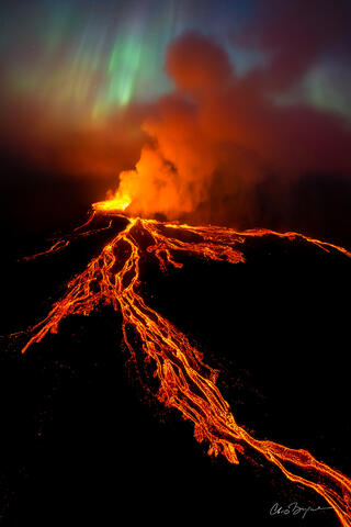 Aerial photograph of a volcano at night shows finger-like orange lava flows, orange molten lava bubbling out of the volcanic crater and northern lights in the s