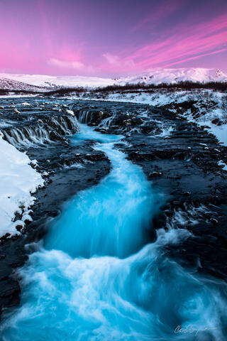 Pink clouds fill the sky behind snow covered mountains as a blue waterfall cascades down black volcanic rock.