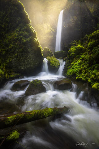 Waterfall enters the gorge with a tall drop where the sun shines in and creates a misty fog while the river tumbles through the rocks below