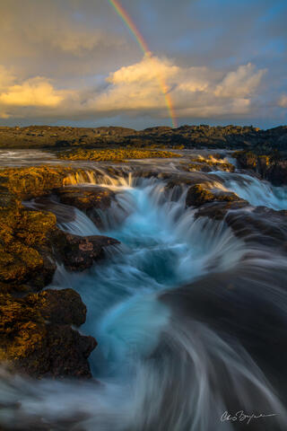 A series of waterfalls along the black volcanic shore of hawaii rush down into wells and back out to sea as a rainbow is on the horizon with sunlit clouds.