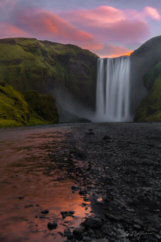 PInk clouds in the sky behind a waterfall over lush green cliffs and the view reflected in the river that flows away from the waterfall.