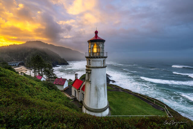 A lighthouse stands on the shore near a sandy beach with a rainbow coming from the distant clouds on the horizon that are lit with sunlight.