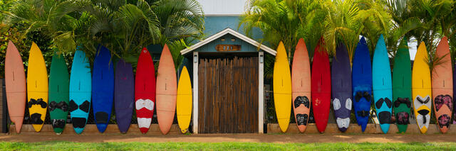 A fence made of colorful surfboards of yellow, red, blue, and green on the sides of a blue roofed surf hostel in Paia on the island of Maui