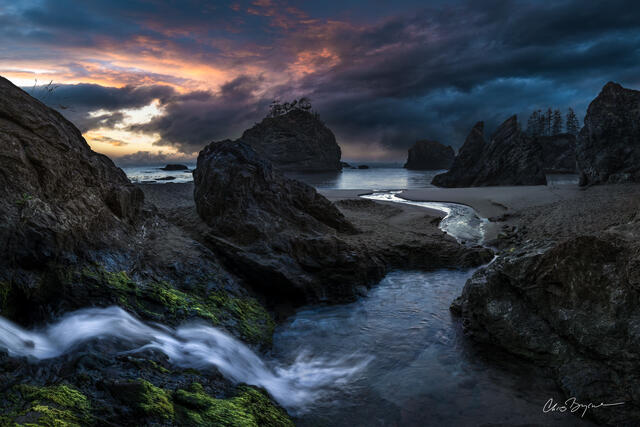 A waterfall flows down to the ocean and the ocean meets the waterfall as the sun sets beneath the dark clouds.