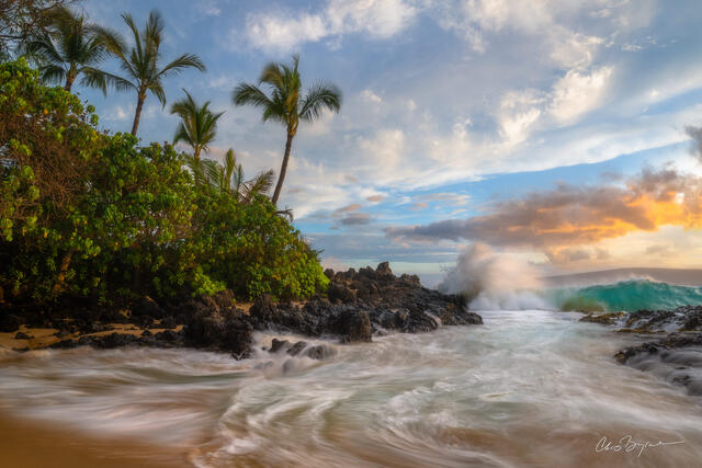 Sunset on a sandy beach with waves washing up on a sandy shore with black volcanic rock up against the lush green palm trees and shrubs that sit back from shore