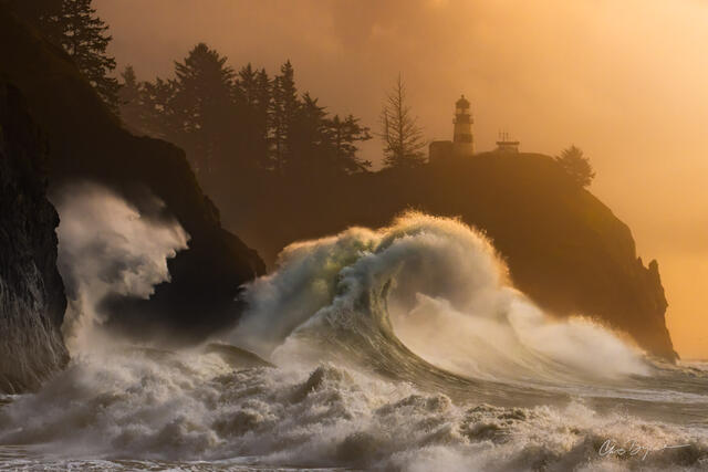 Large ocean waves crash into the rocky coast beneath a lighthouse that sits on a cliff at sunset with an orange glow in the sky.