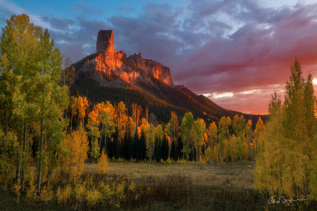 Yellow aspen trees and a mountain topped with a chimney-shaped rock are lit up by the sunset as a orange and pink sky glows across the scene.