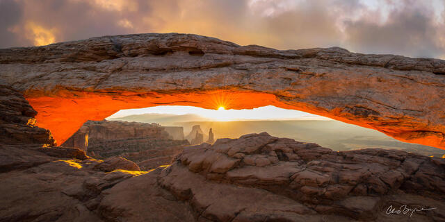 The sunrise lights up the underside of a sandstone arch that shows the canyon below and mountains in the distance.