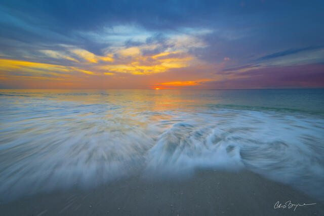 The white water meets the beach as the sun sets and lights the sky and water with pastel colors from the blue water to the pink skies and clouds.