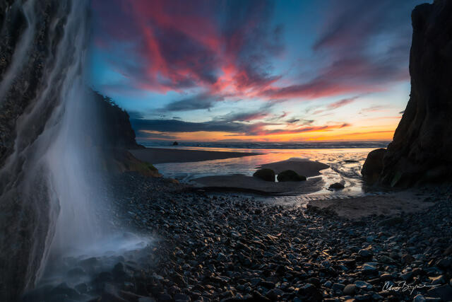 The edge of a waterfall is barely in view that looks out to the ocean where the sun sets along the coastline and lights the sky with pink and orange clouds.