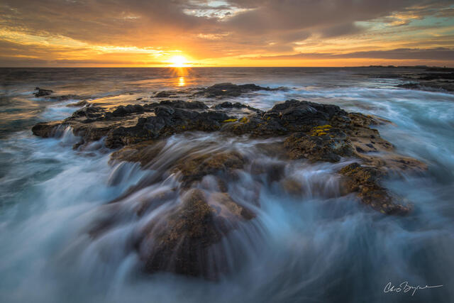 Along the volcanic rocky shore of hawaii water rushes in and cascades over the black rock while the sun sets on the horizon shining its rays and reflecting on t