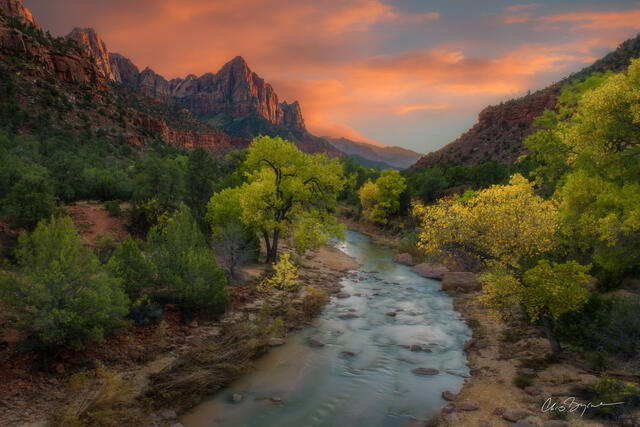 A river flows through a canyon as the sun rises and lights up the large rock formation and sky with pink clouds.
