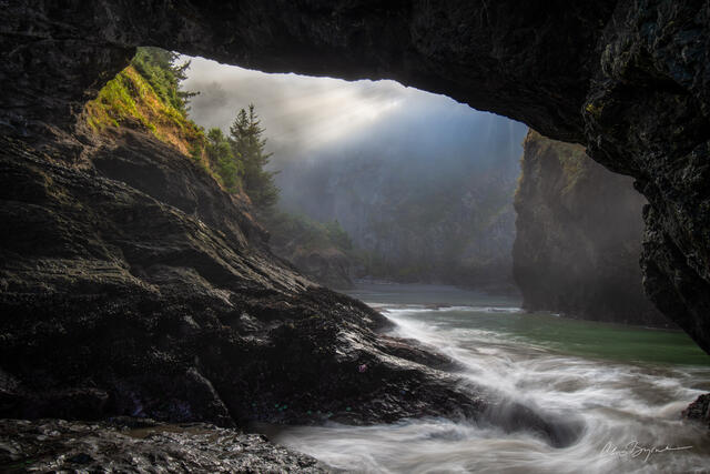 A cove surrounded by dark rock looking out as sun rays shine through the mist and water rushes into the cove while spruce trees are seen on the river banks.