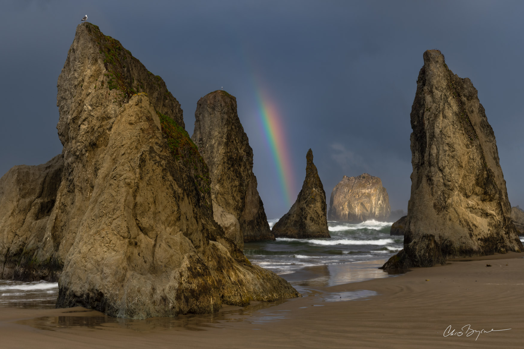 PIRATE SHIPS Sea stacks on the Oregon coast surrounded by ocean waves and darky clouds in the sky with a rainbow in the distance.