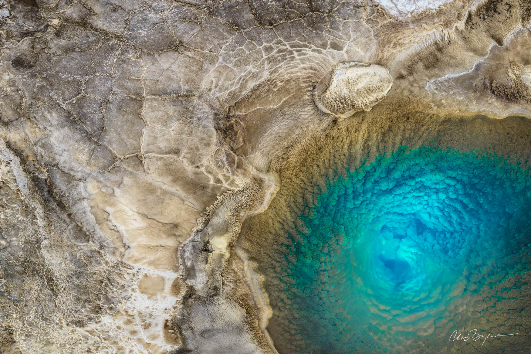 UNDERWORLD Aerial photograph of a geyser filled with bright blue water and surrounded by light tan rock creates an abstract image of what looks like an eye.
