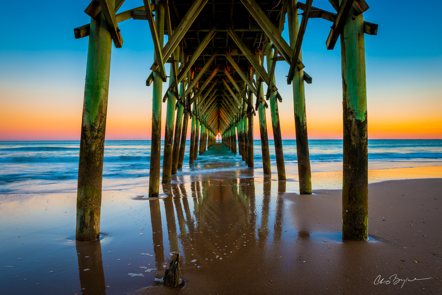UNDER THE PIER IN WINTER A tall wooden pier seen from underneath as the water rushes in to the sandy shore and the sun sets on the horizon.