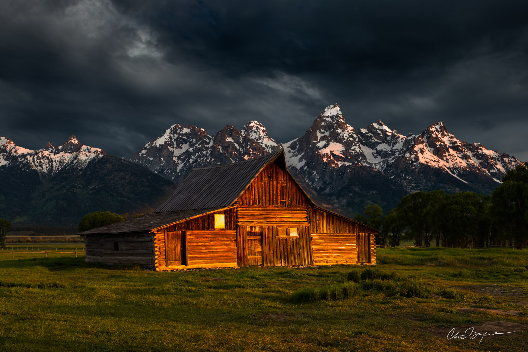 HOME ON THE RANGE Sun shines on the front side of a rustic barn that sits in front of grand mountains with snow covered tops and dark clouds in the sky.